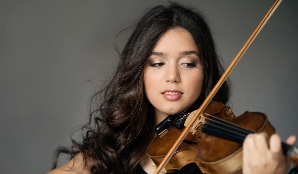 A young women with dark hair, looking down at her violin.